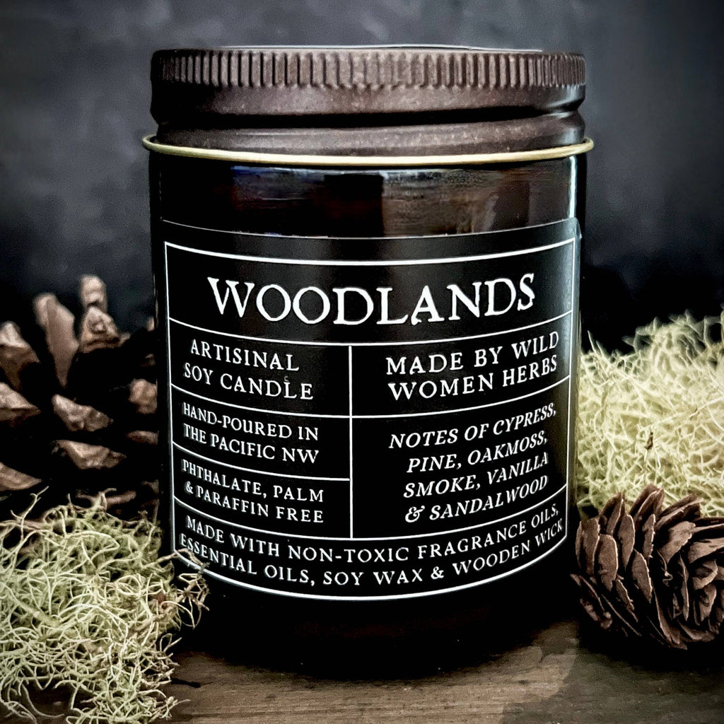 Candle labeled 'Woodlands' by Wild Women Herbs on a wooden surface with natural elements.
