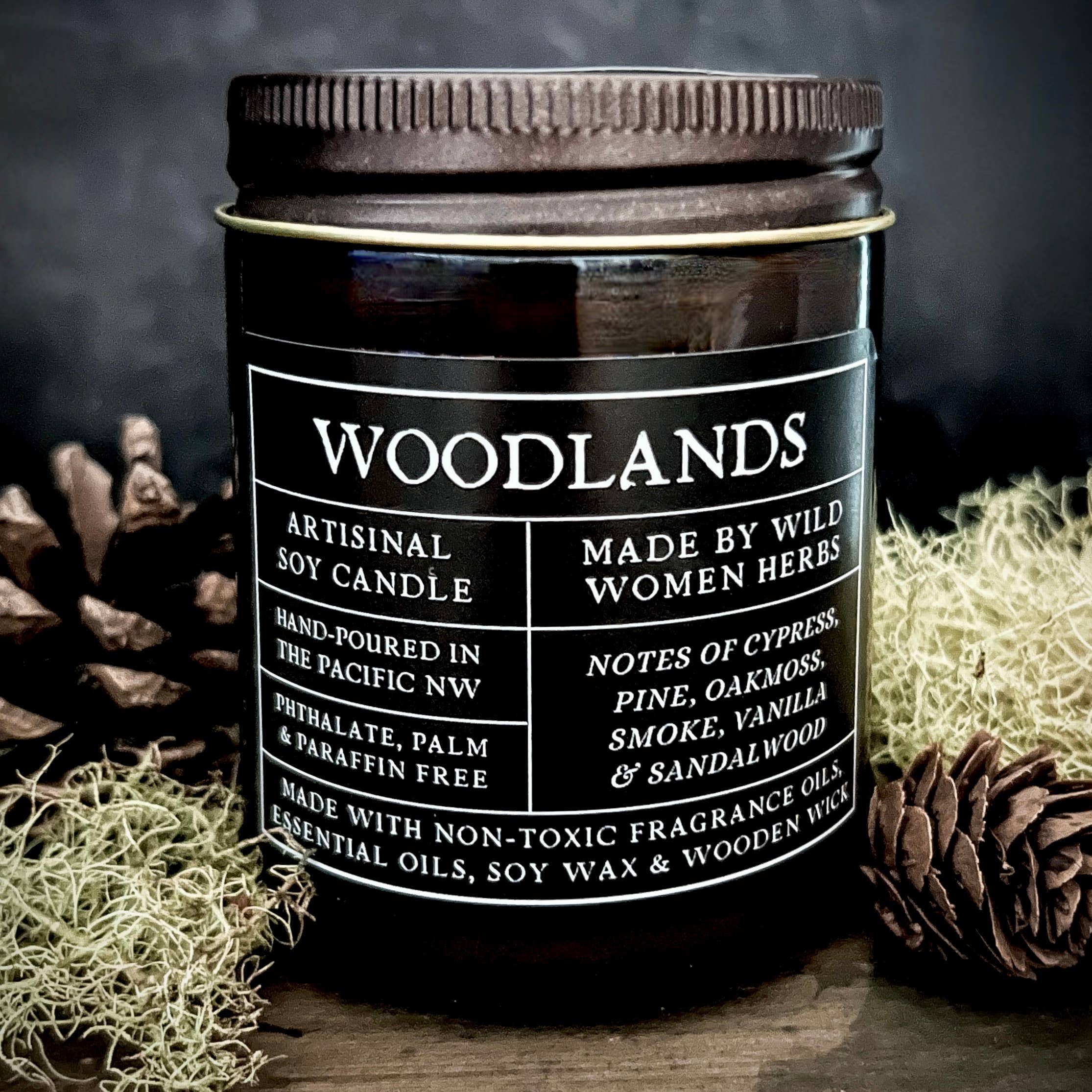 Candle labeled 'Woodlands' by Wild Women Herbs on a wooden surface with natural elements.