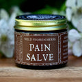 A small jar of Wild Women Herbs Pain Salve sits on a rustic wooden surface, framed by white flowers and green leaves against a dark, blurred background.