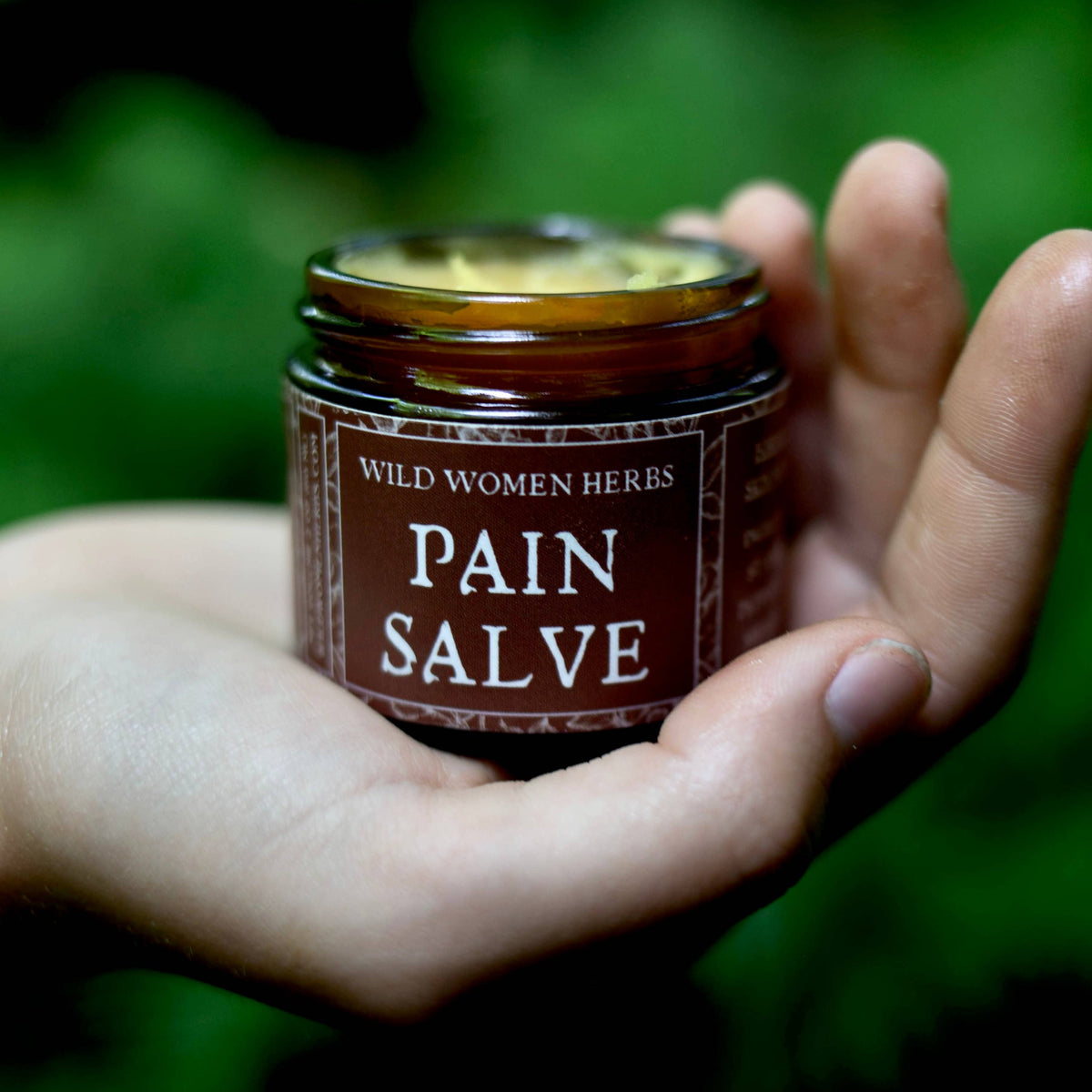 A hand holding a small amber glass jar of Wild Women Herbs Pain Salve against a blurred green outdoor background.