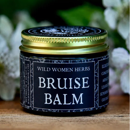 A small tin of Wild Women Herbs Bruise Balm sits on a rustic wooden surface, surrounded by soft-focus white flowers and green leaves.