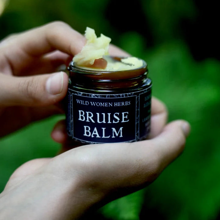 A person's hands hold an open amber glass jar of Wild Women Herbs Bruise Balm against a blurred green outdoor background, with some balm visible on top of the jar.