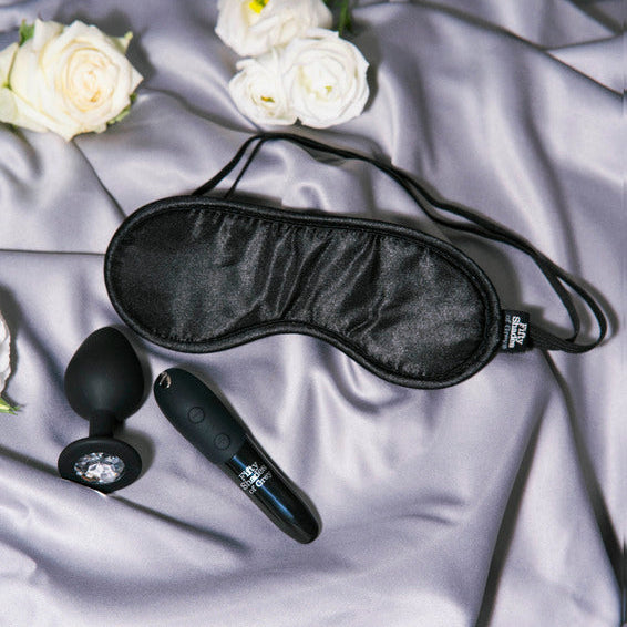 A bullet vibe, blindfold and plug et on a satin sheet with flowers around them.