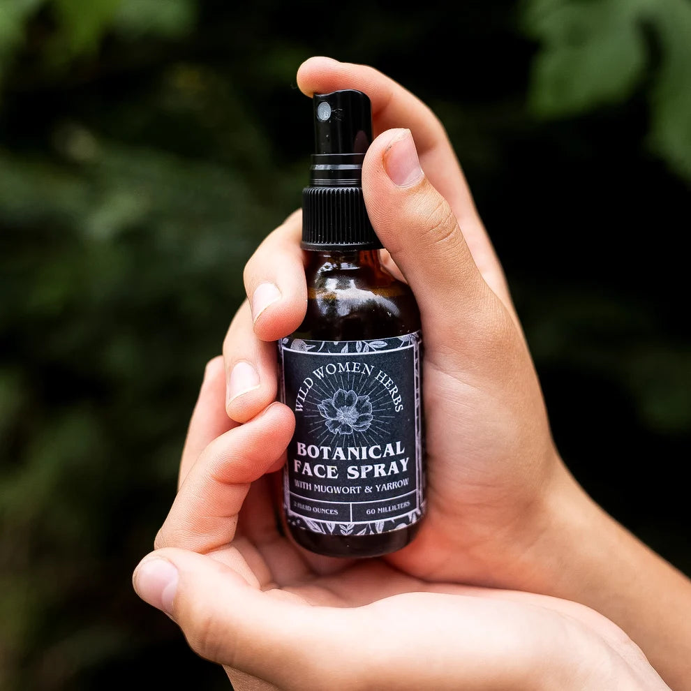 A person's hands holding a small amber glass spray bottle labeled 'Wild Women Herbs Botanical Face Spray' against a blurred dark green outdoor background.