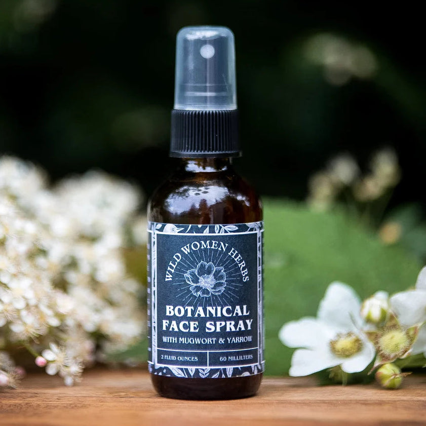 A bottle of Wild Women Herbs Botanical Face Spray stands on a wooden surface with white flowers and green foliage in the background.