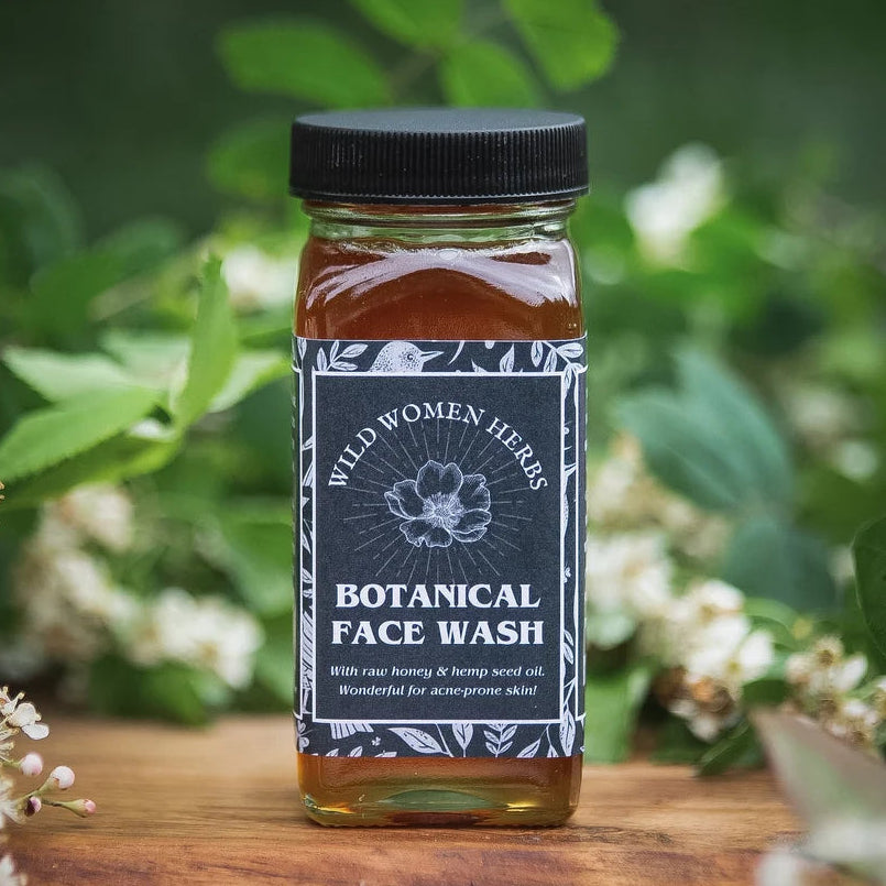 A bottle of Wild Women Herbs Botanical Face Wash stands on a wooden surface surrounded by green leaves and white flowers. The bottle contains an amber-colored liquid and has a black cap with a dark gray label featuring white text and botanical illustrations.