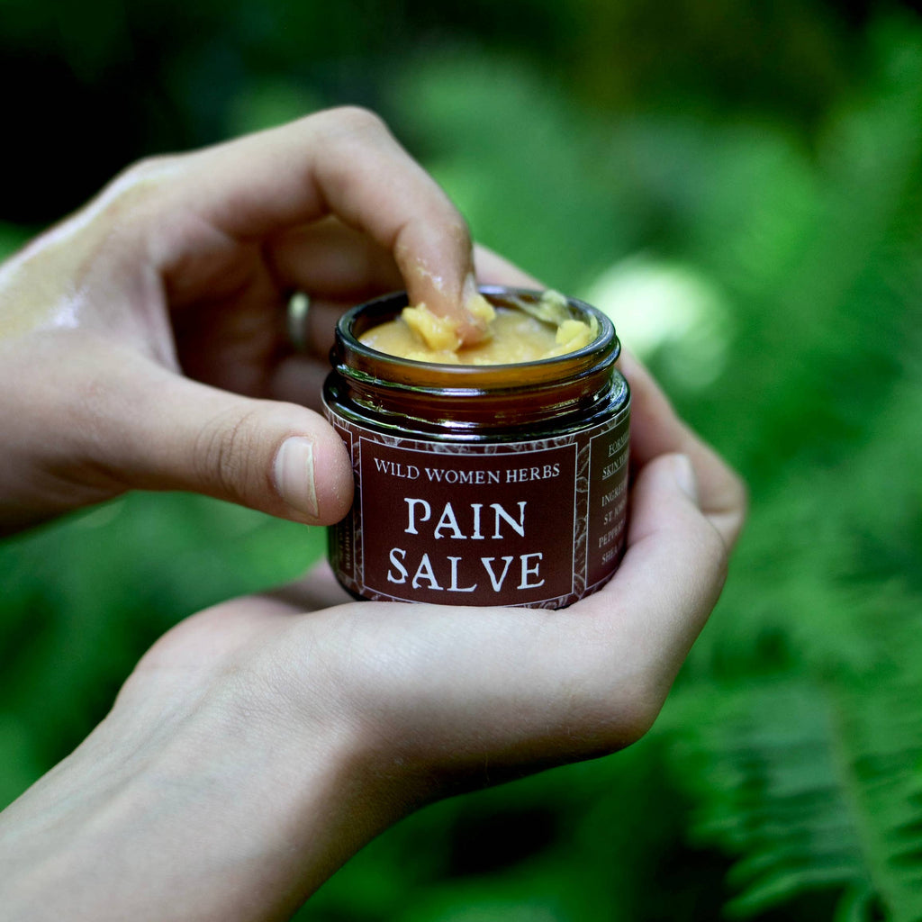 A person's hands holding a small amber glass jar of Wild Women Herbs Pain Salve, with a piece of salve being spread from the top, set against a blurred green fern forest background.