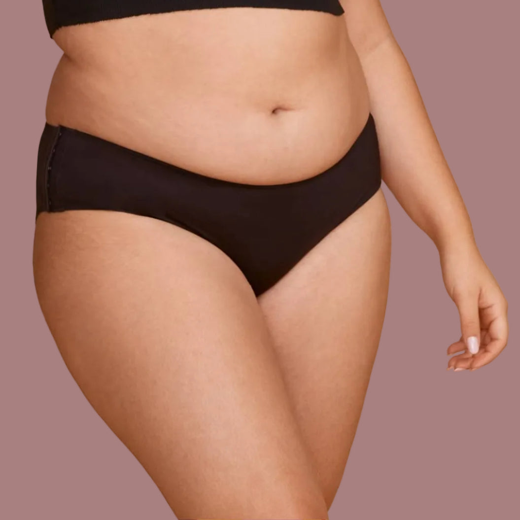A woman modeling black classic hipster panties, photographed against a plain light beige studio background.