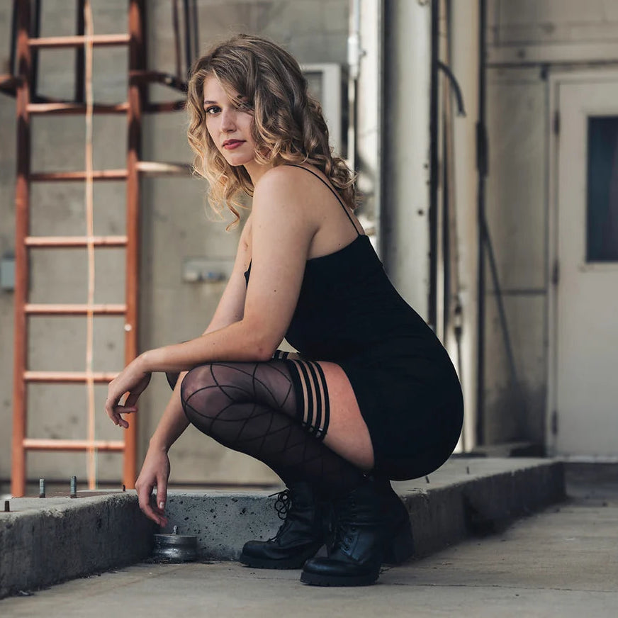 Woman in a black outfit squatting in an industrial setting while wearing diamond knit thigh high stockings.