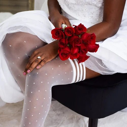 A close-up lifestyle shot of a bride sitting on a black chair, wearing sheer white tights with small white polka dots and white thigh-high stockings with three stripes, holding a bouquet of red roses.