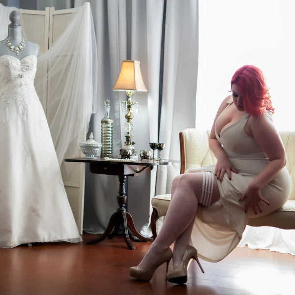 Woman sitting on a couch next to a wedding dress in a room with curtains and a lamp.