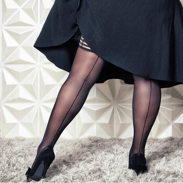 Woman in a black coat standing against a geometric-patterned wall while lifting her skirt to show the top of her seamed stockings.