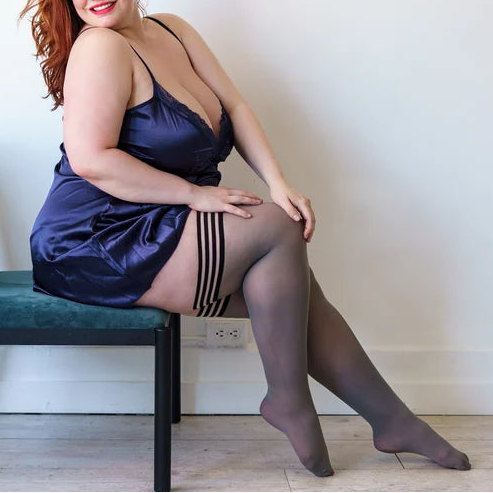 Woman sitting on a chair wearing a satin dress and stockings in a room.