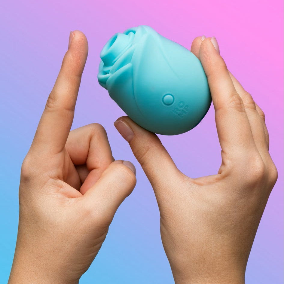 A light blue silicone personal massager shaped like a stylized rose, held between two hands against a pink and blue gradient background.
