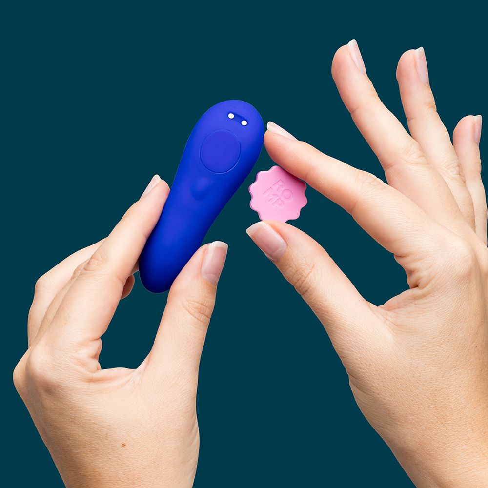 A close-up of a person's hands holding a blue personal massager and a small pink flower-shaped magnet against a solid dark teal background.
