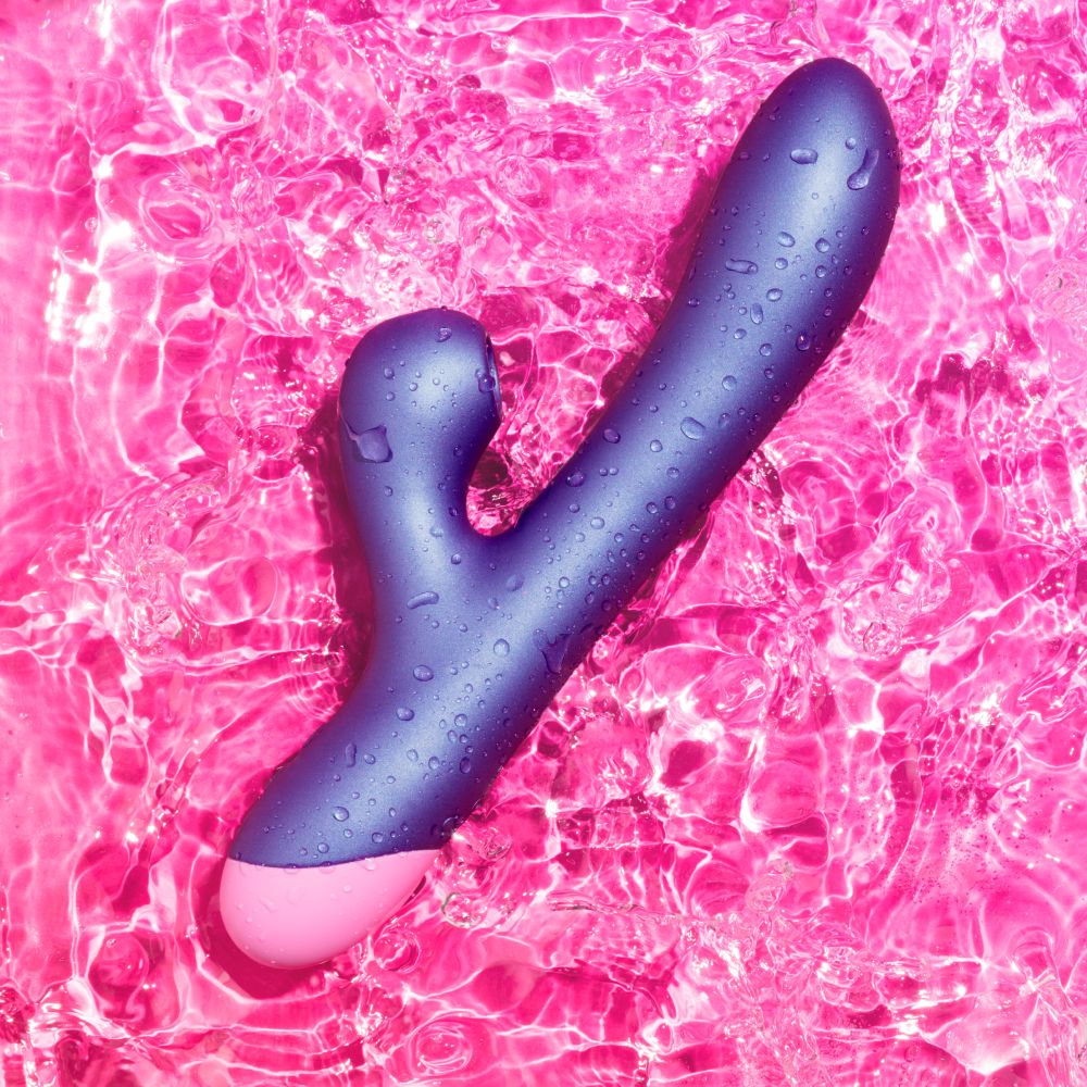A personal massager with a matte dark purple body and a pink base, covered in water droplets and set against a vibrant pink swimming pool background.