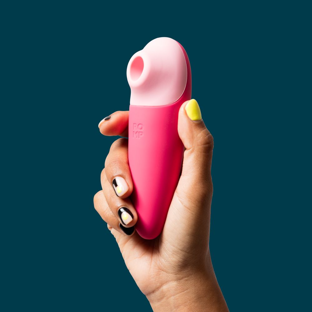 A hand with decorated nails holds a pink and light pink personal massager against a solid dark teal background.